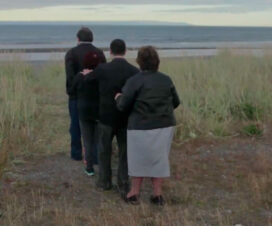 Captura de pantalla de una escena del cortometraje Los Bastones del Actor. Tres personas de espaldas, caminan en fila tomados por los hombros por un campo.