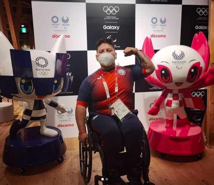 Marion Serrano levanta su brazo izquierdo mostrando sus músculos sentada en su silla de ruedas, a cada lado suyo están hay figuras de las mascotas de los Juegos Olímpicos y Paralímpicos de Tokio 2020.